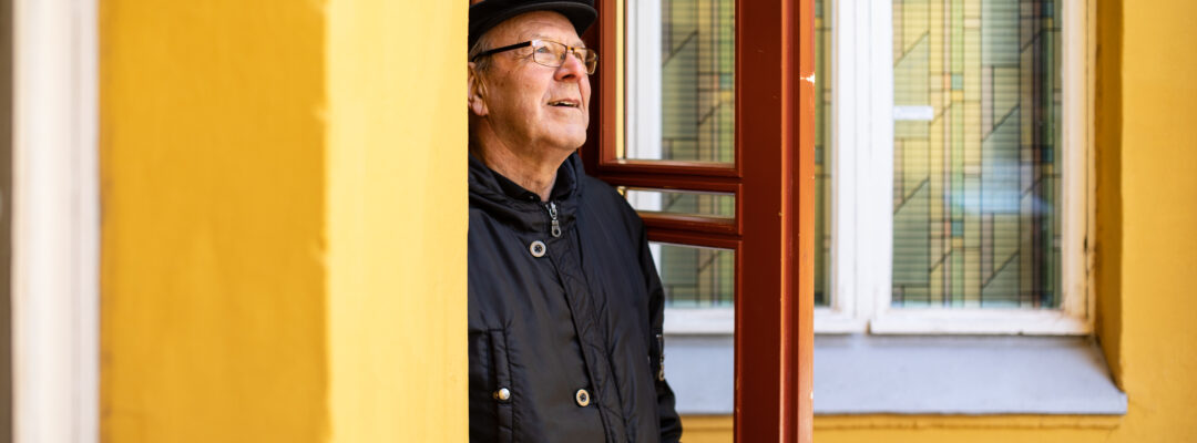 Eläkeläinen avaa oven. Rakennus on keltainen ja ovi ruskea.
