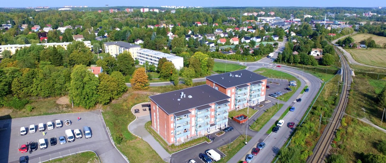 Flygbild över bostaden på Raunistulavägen. I förgrunden finns tågbanan och vägen som leder till fastigheten och själva fastigheten som är ett höghus i fyra våningar. Det finns mycket parkeringsplats utanför huset. I bakgrunden finns Raunistula och dess bostadsområdet med småhus och höghus.