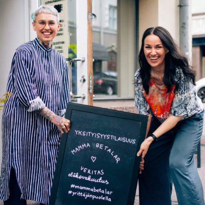 Tilannekuva, jossa vaikuttajat Nata Salmela ja Jasmin Hamid hymyilevät iloisesti ja pitelevät Veritaksen ja Mamma Betalar -podcastin yhteistyötilaisuuden kylttiä kädessään.