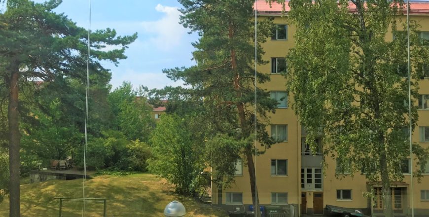 Helsingin Meripuistotien asunnon parvekkeen näkymää kesäisenä päivänä. Pihalla on nurmialuetta ja muutama korkea puu. Vastapäätä on keltainen kuusikerroksinen kerrostalo.