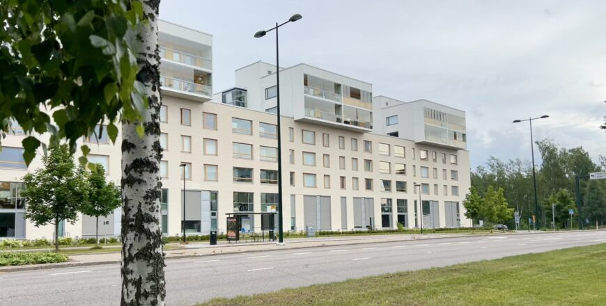 Nuttukujan kiinteistö etäältä kuvattuna. Rakennuksen edessä on puita ja autotie. Rakennus on väriltään vaalea.