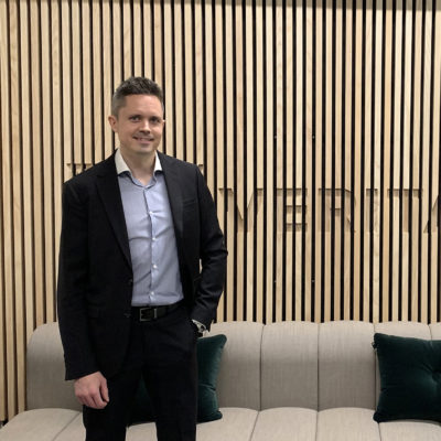 Jussi häkkinen seisoo puisen seinän edessä, jossa lukee "Veritas". Taustalla on myös harmaa sohva.