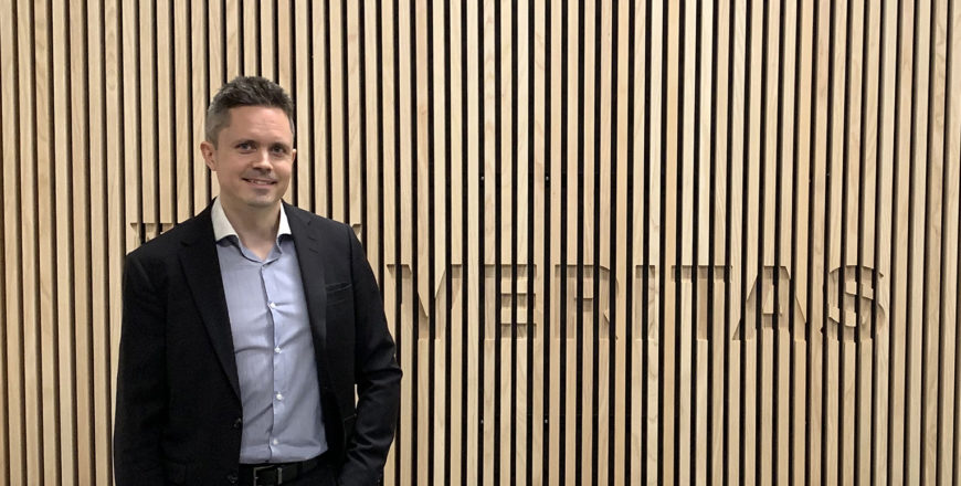 Jussi häkkinen seisoo puisen seinän edessä, jossa lukee "Veritas". Taustalla on myös harmaa sohva.