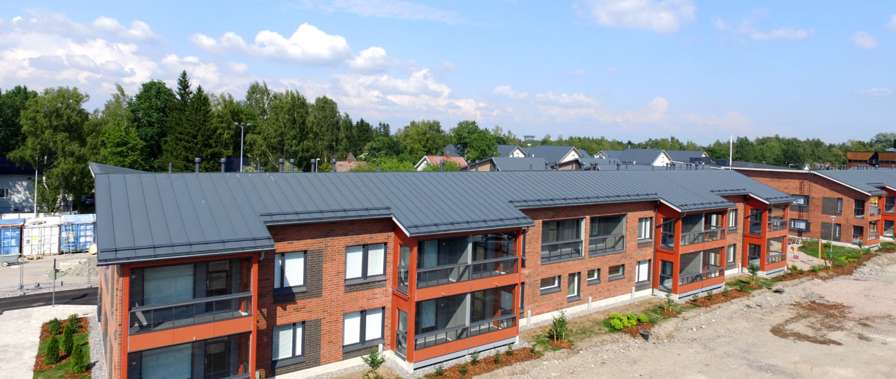 Fasaden på Vantaan Kuunliljas hyreslägenheter. Huset har röd tegelfasad och svart plåttak. Lägenheterna ligger på två våningar och balkoger är inriktade mot innergården. Mellan husen finns en lekplats.