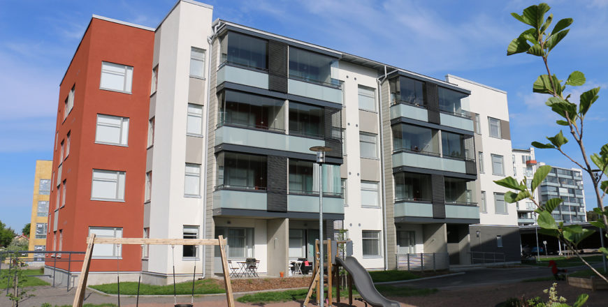 Fasaden på Pryssgatans höghus. Huset är rödbrunt och vitt och har fyra våningar och inglasade balkonger. På gården finns lekplats för barn och torkställning.