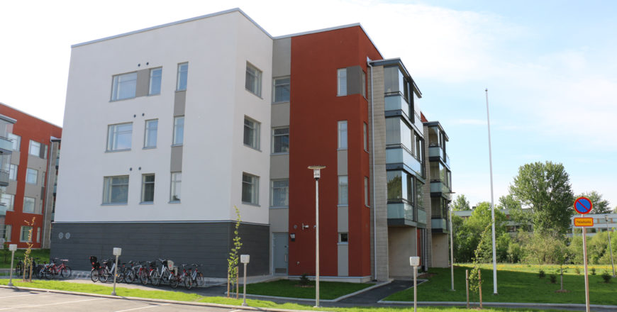 Facaden på Pryssgatans lägenhet från sidan. Huset är i fyra våningar och är vitt. På gården finns parkeringsplatser och lekplats för barn.