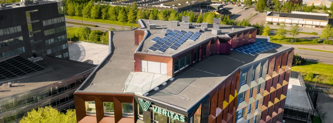 Kesäinen ilmakuva Veritaksen värikkäästä pääkonttorista, Retoriikasta, jonka katolla on kymmeniä aurinkopaneeleja. Taustalla näkyy Helsingin valtatie ja sen takana olevia muita kiinteistöjä.