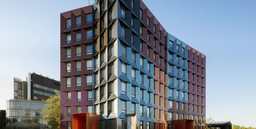 Veritaksen pääkonttori Trivium Retoriikka kuvattuna etupihan parkkipaikalta. Rakennus on yhdeksänkerroksinen ja monivärinen. Rakennuksen sivulla on pieni istutusalue sekä pyöräkatos.