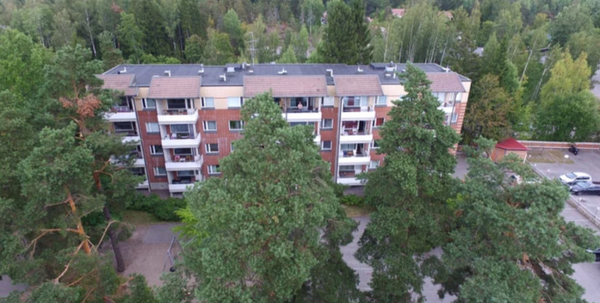 Ruutikellarintien kerrostalon ilmakuva, josta näkyy ympärillä olevaa metsäistä maisemaa. Pihalla on tuuletusteline, leikkipaikka sekä reilusti parkkipaikkoja.