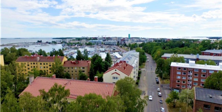 Ilmakuva Meripuistotien asuntojen lähialueesta. Etualalla on muita taloja sekä autotie, jonka molemmin puolin kulkee kävelytiet. Kauempana on näkyvissä rantaa, venesatamaa ja Helsingin Lauttasaari.