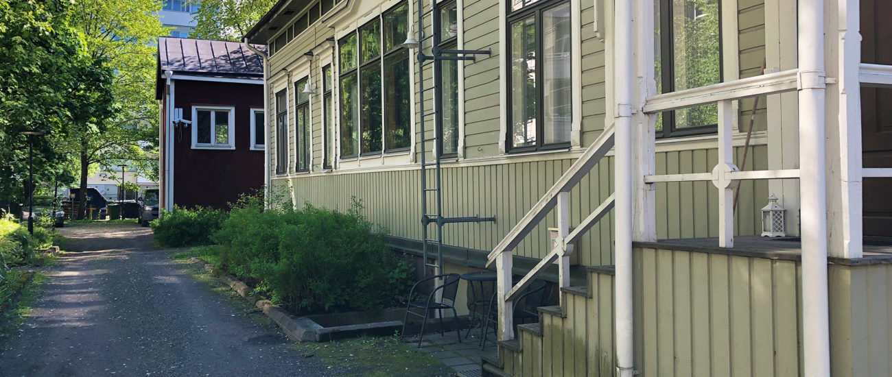 Linnankadun puutalo sisäpihan puolelta kuvattuna. Pihalla on hiekkatie ja kesäisen vehreitä puita sekä pensaita. Asuntojen sisäänkäynnissä on useampi porras.