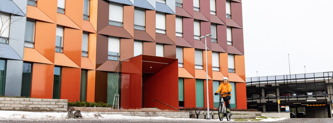 Henkilö ajaa sähköpyörällä Turussa Veritaksen pääkonttorin edessä. Rakennus on värikäs ja maassa on vähän lunta. Taustalla näkyy parkkihalli.